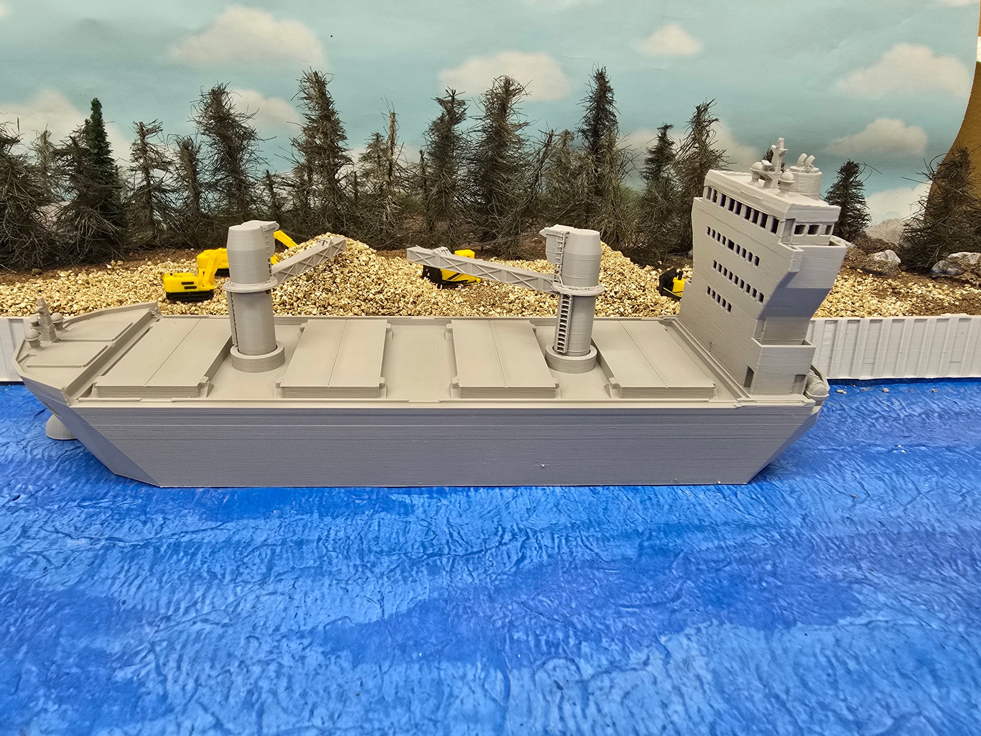 Model of a large ship on a blue surface with trees and construction vehicles in the background