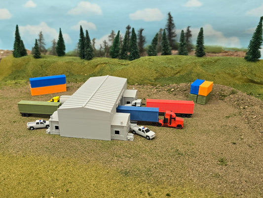 Model of a warehouse with colorful shipping containers and vehicles on a grassy field with trees in the background.