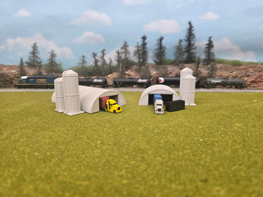 Model train scene with tunnels, trucks, and trains on a grassy field.