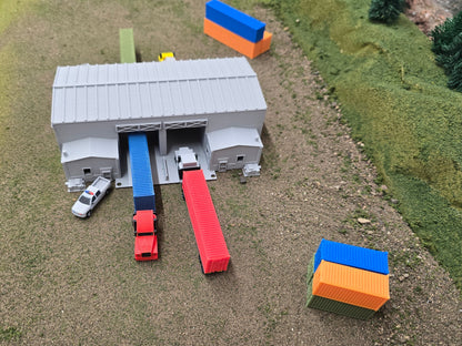 Model of a train station with a gray building, red and blue train cars, and a white car on a grassy field.