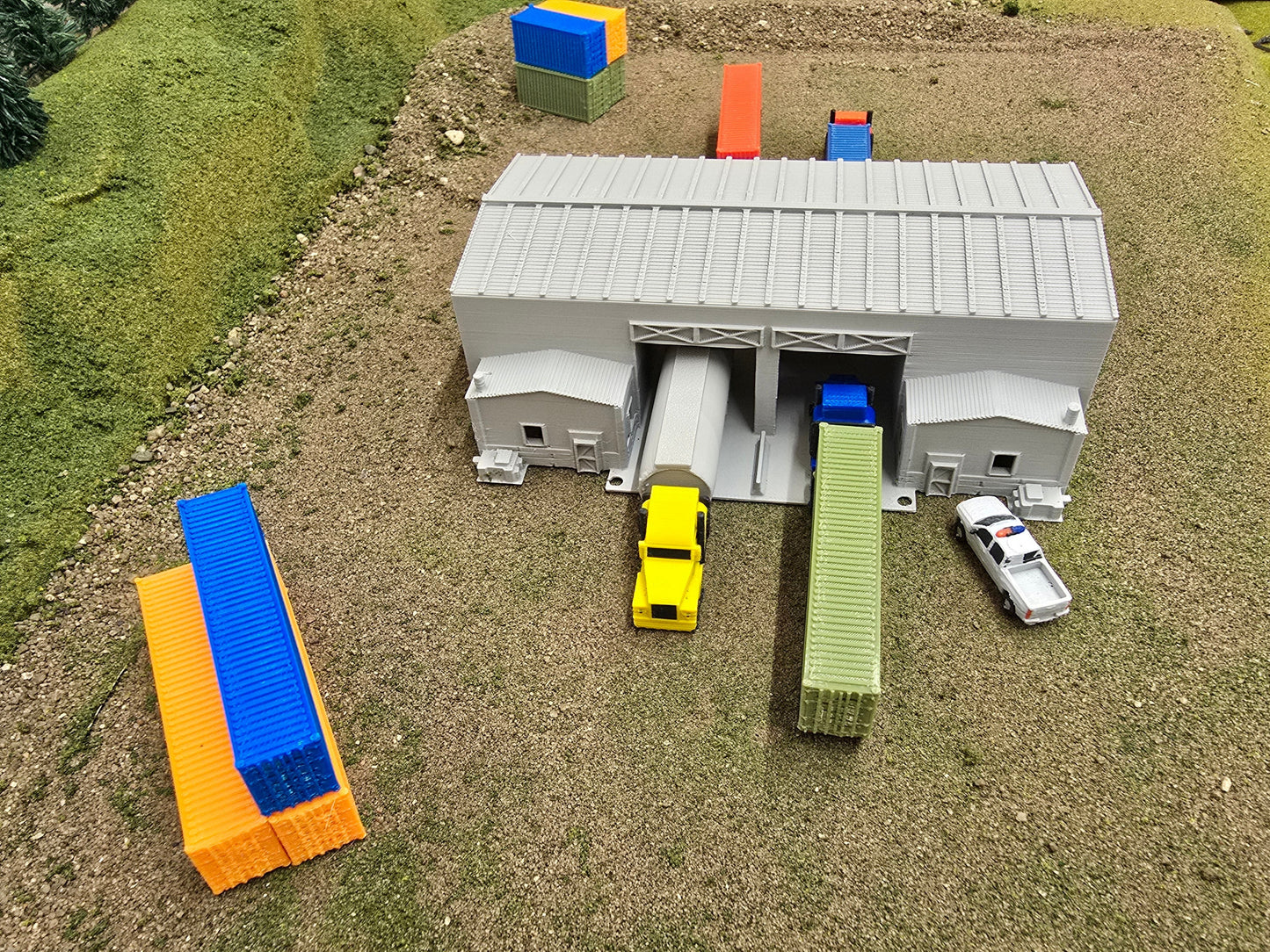 Model train layout with a gray building, yellow truck, and other miniature vehicles on a grassy surface.